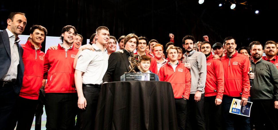 Photo: A large group of hockey players surround Macklin Celebrini who is standing behind the 2024 Hobey Baker Award trophy