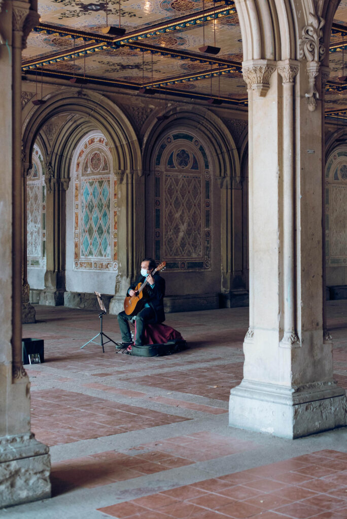 Photo: A lone man sits and plays a cello with a mask on in a grand area with tall pillars and archways.
