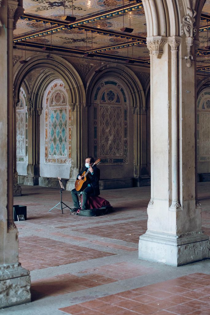 Photo: A lone man sits and plays a cello with a mask on in a grand area with tall pillars and archways.