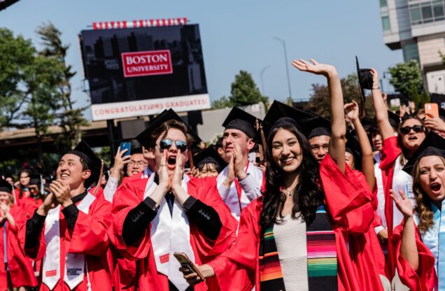 A celebratory scene at Boston University with a group of graduates in red robes and black caps, joyfully raising their hands and capturing photos under a clear blue sky. They are standing in front of a large billboard that reads ‘BOSTON UNIVERSITY CONGRATULATIONS GRADUATES!’ in bold white letters on a red background, marking the festive occasion of their graduation.