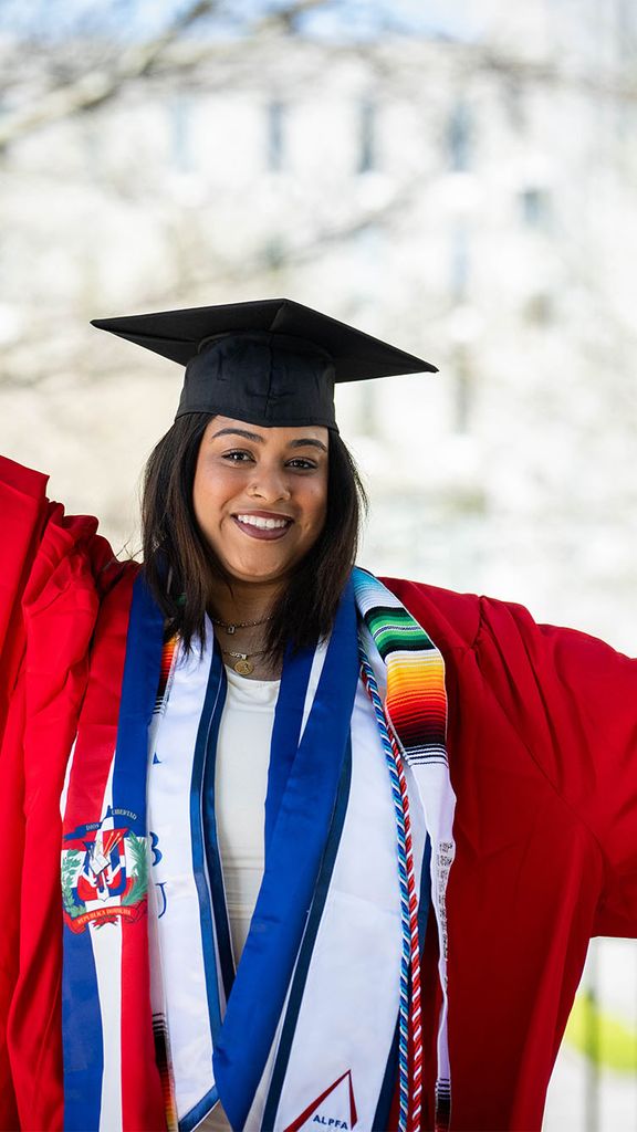 Photo: A college student in a red robe with black mortarboard and blue tassles holds their arms up in celebration