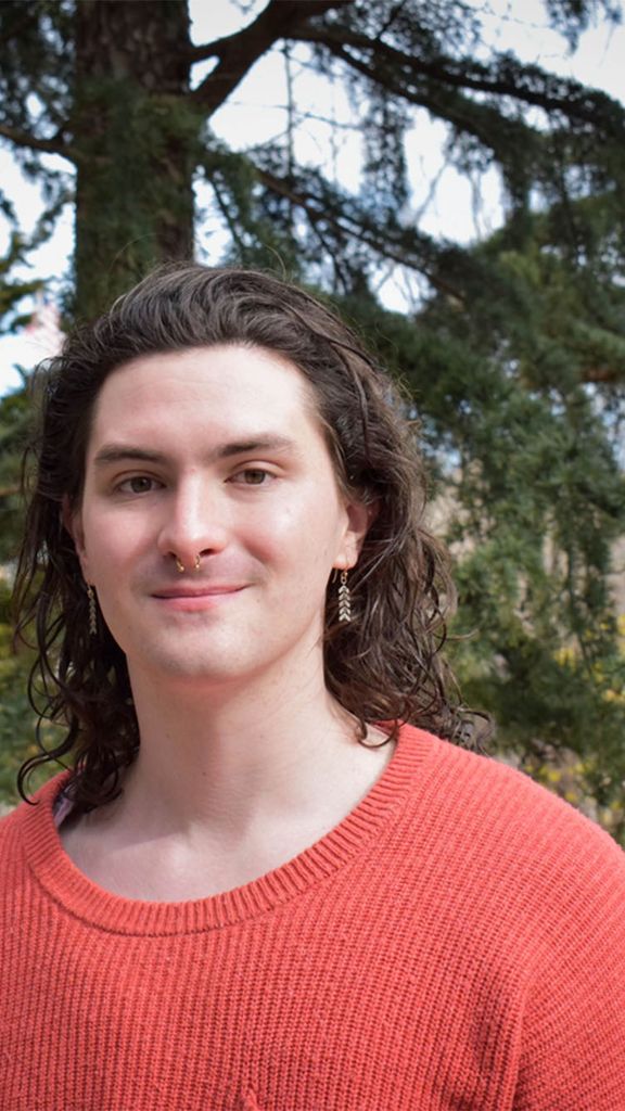 Photo: A person with short dark hair and a nose ring wearing an orange shirt in front of a verdant nature scene with pine trees