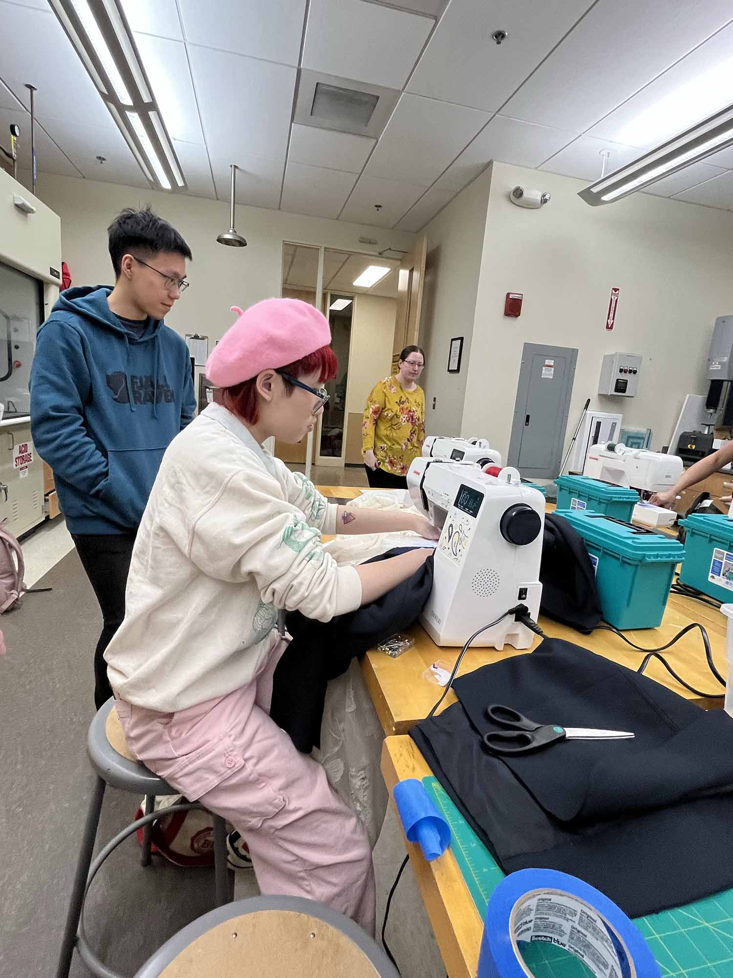 Photo: Kal Hawley, an individual with red hair, pink beret and pants (left), sewing lace on their blazer as group member Sky Lan (CAS’25) looks on.