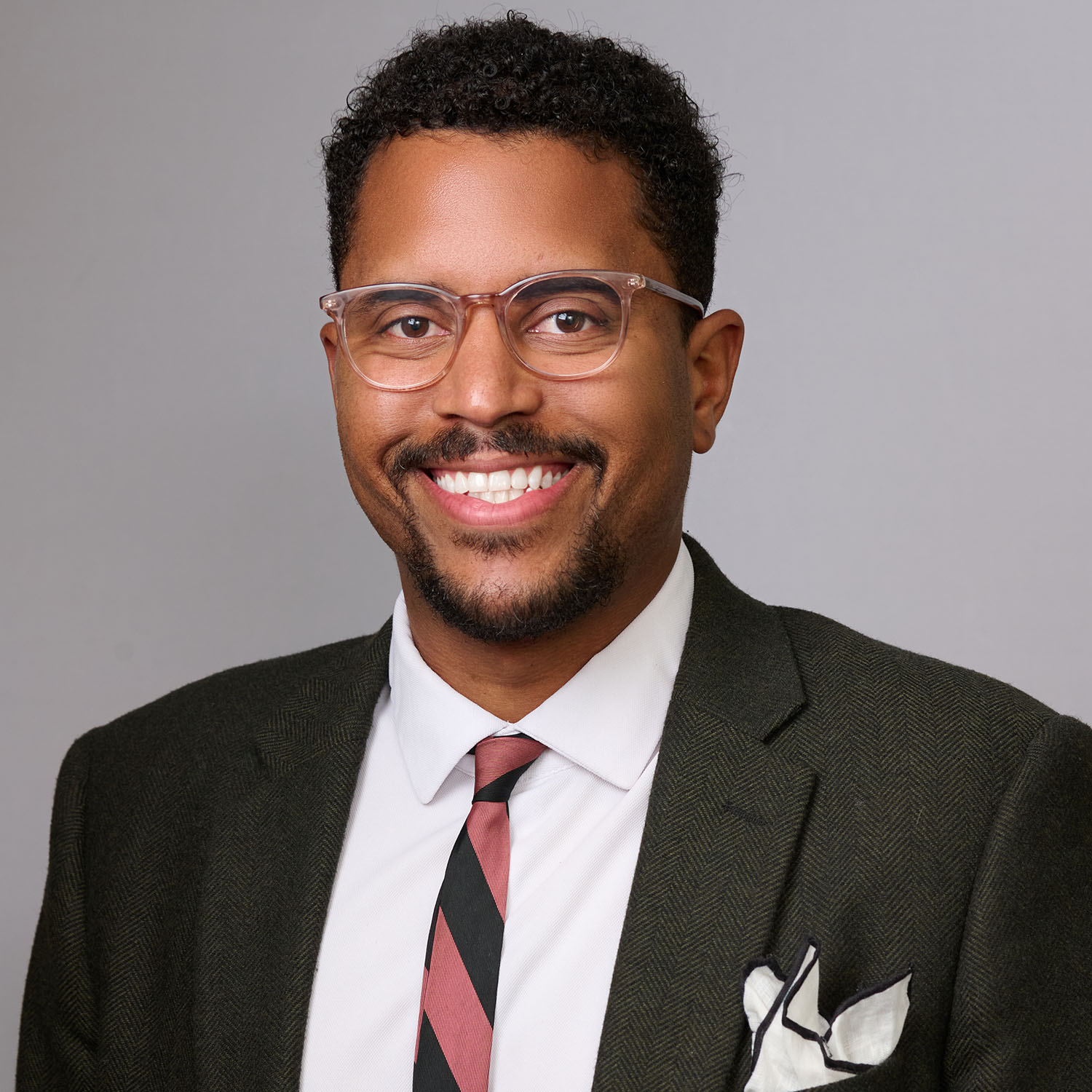 Photo: A headshot of a man with dark hair and a beard smiling. He is wearing clear-framed glasses and a dark suit