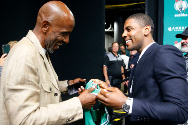 Photo: two black men in formal dress exchange a green basketball jersey with smiles on their faces. In 2017, Kyrie Irving gifted his father Drederick (CAS’88) his first Boston Celtics jersey after a news conference in Boston. Kyrie played for Boston from 2017 to 2019. Photo by Winslow Townson via AP