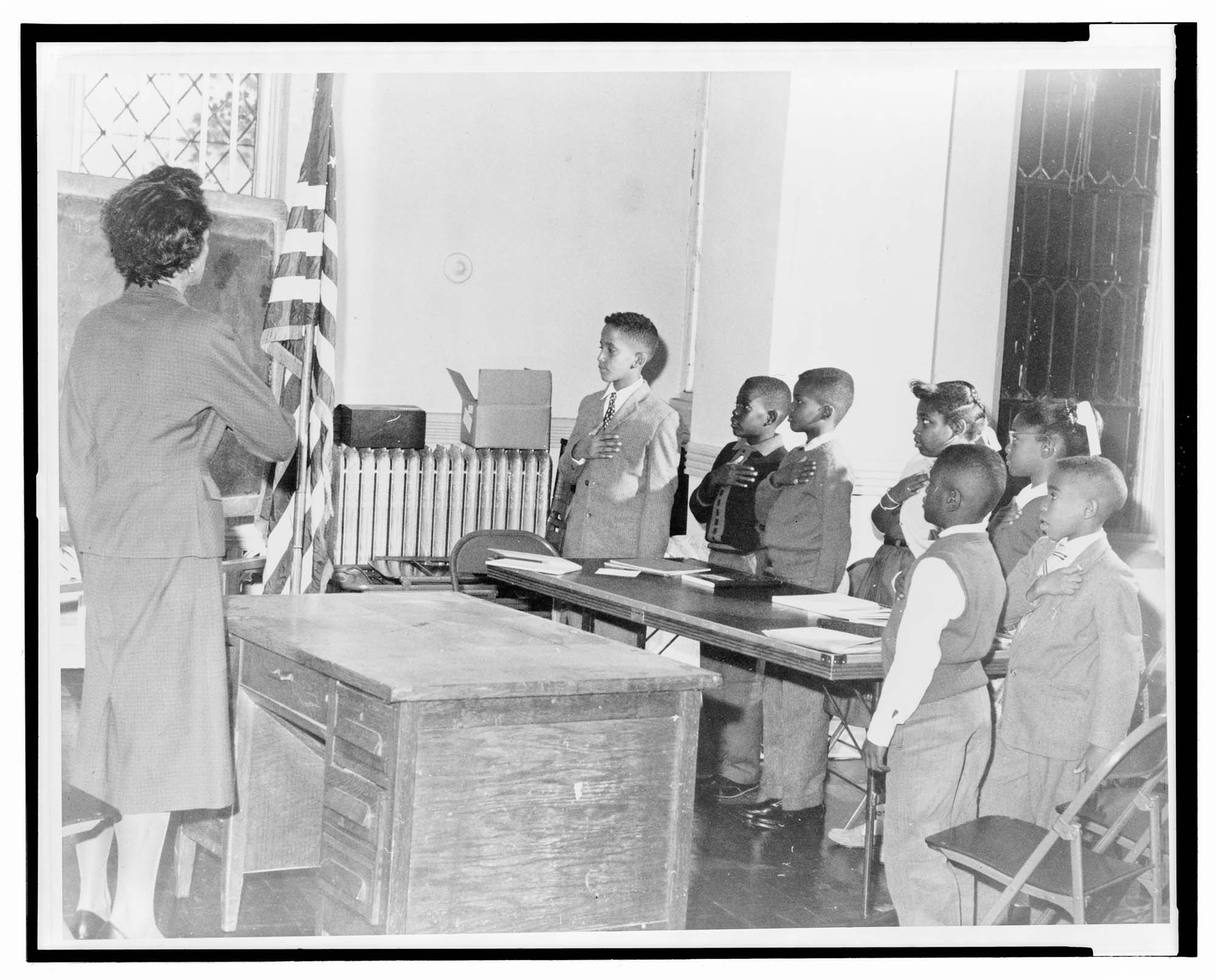 Black and White photo of a woman teaching a class of african american school children. Everyone is dressed in attire from the 1960s as they salute the American flag