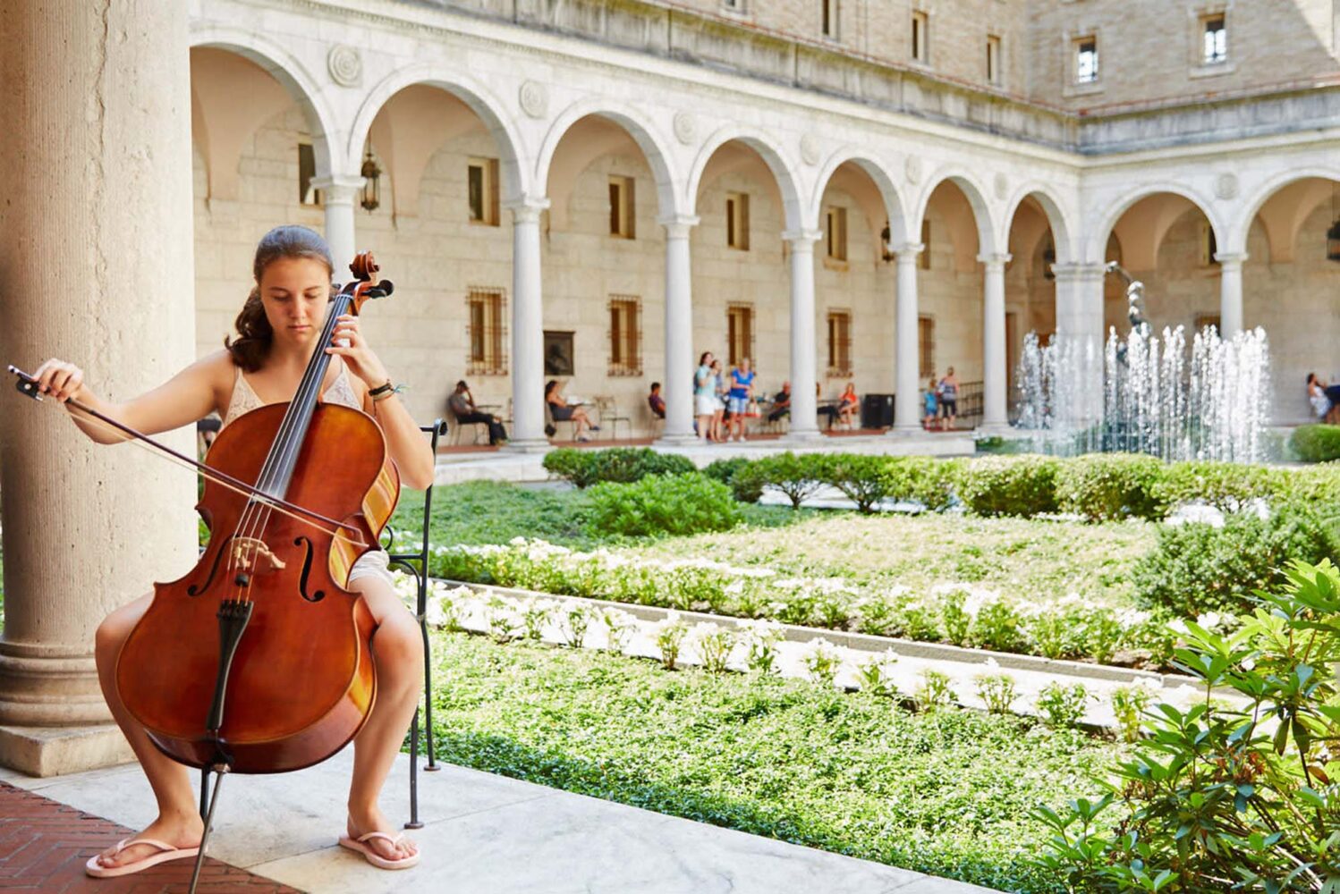 Photo: A picture of a woman playing the cello outside in a courtyard surrounded by greenery with a fountain in the background