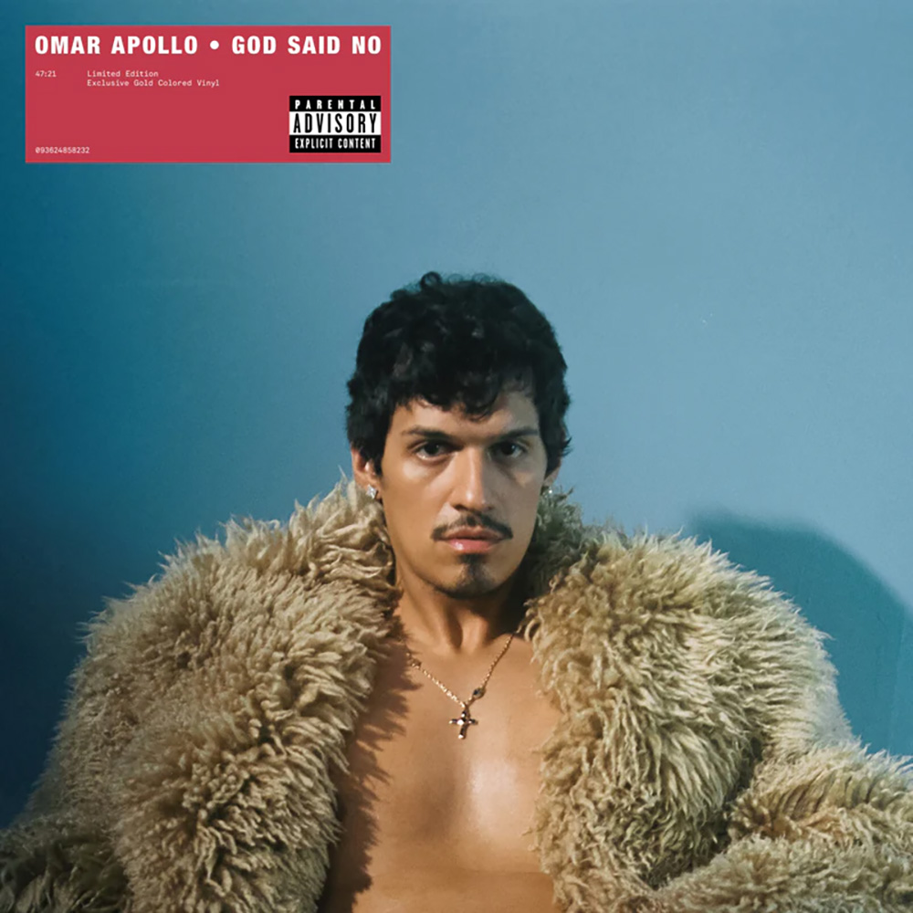 Photo: A portrait of a man, singer Omar Apollo, sitting in front of a blue wall wearing only a fluffy fur coat as he stares down the camera.