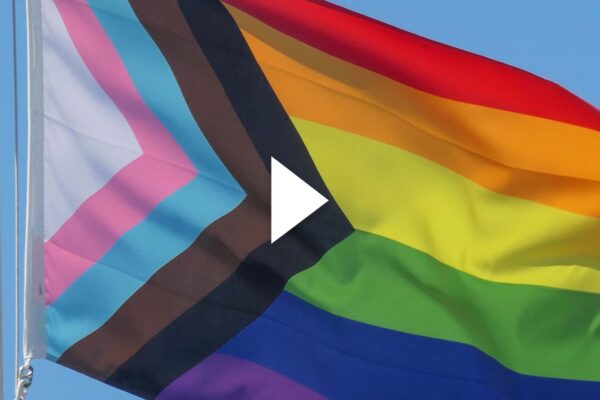 Photo: A picture of a pride flag being raised in front of a blue sky. Play button overlay.