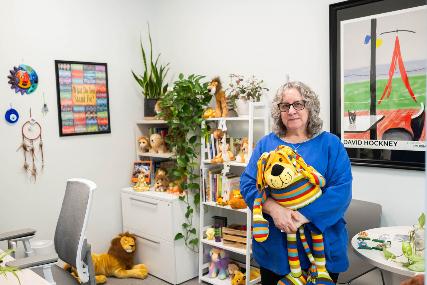 Photo: Roz Abukasis, a women wearing a bright blue shirt and holding a stuffed lion, smiles for a portrait inside her office. Abukasis is an academic counselor at Sargent.