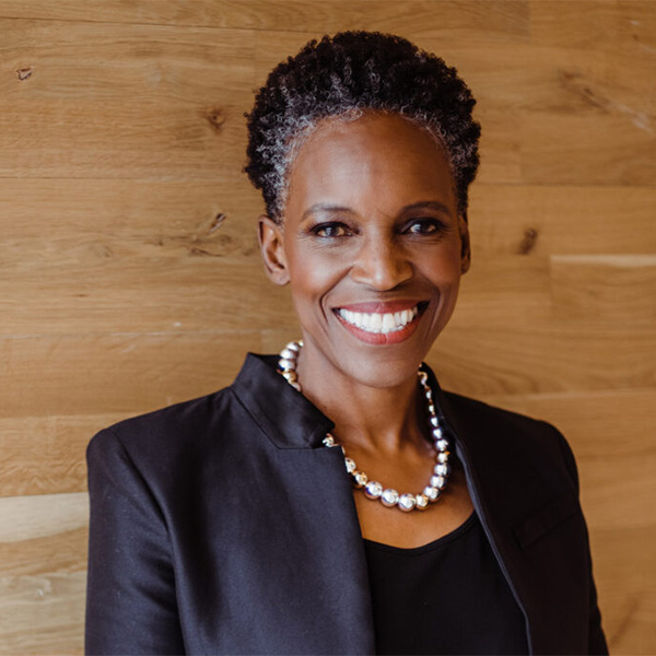 Photo: A Black woman with a short grey and black afro and wearing a necklace of large, faux metal pearls, black blouse, and black blazer, smiles and poses with arms crossed in front of a wodden backdrop.
