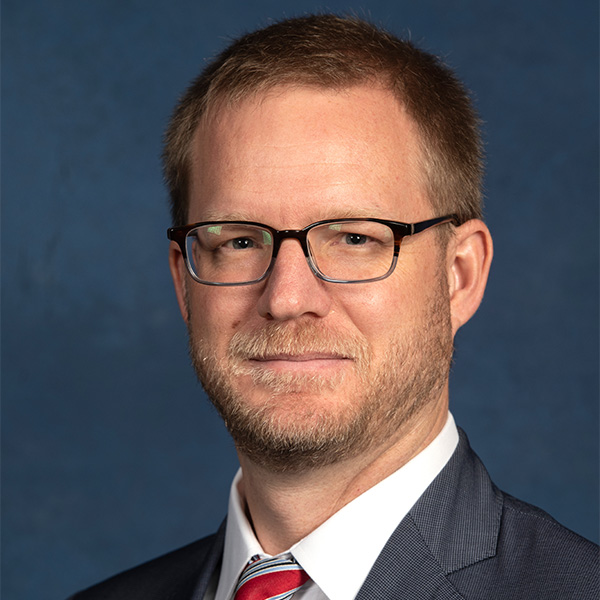 Photo: A white man with short hair wearing a suit and glasses poses for a formal portrait 