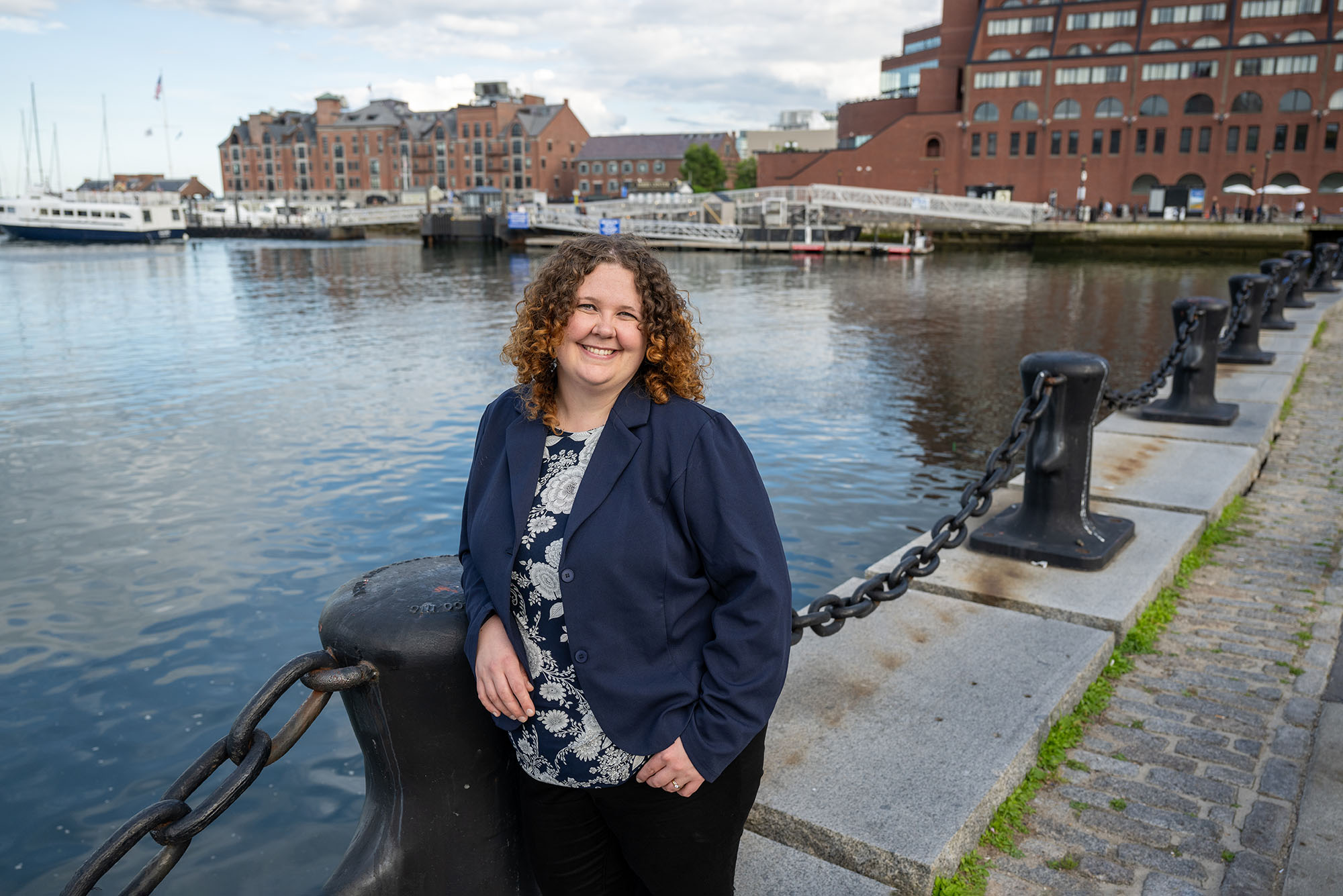 Photo: A picture of a woman with curly brown hair wearing a blue sweater and floral shirt smiling and posing in front of the Boston harbor.