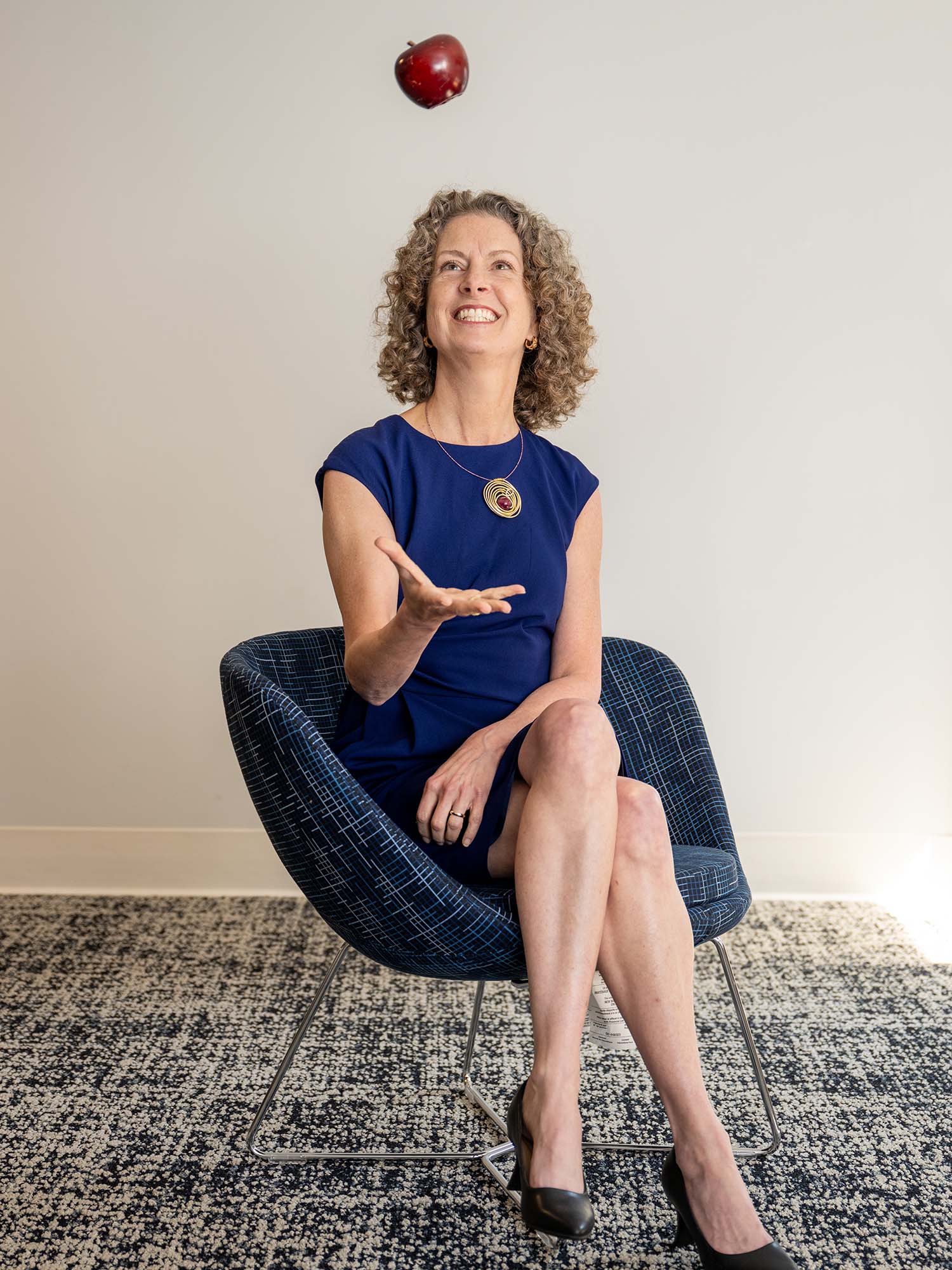 Photo: A picture of Penny Bishop, who has curly short hair and is wearing a blue dress, sitting on a chair and tossing an apple in the air