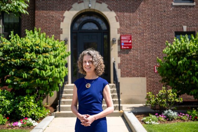 Photo: A picture of a woman with short, curly hair smiling in front of a BU building while wearing a blue dress