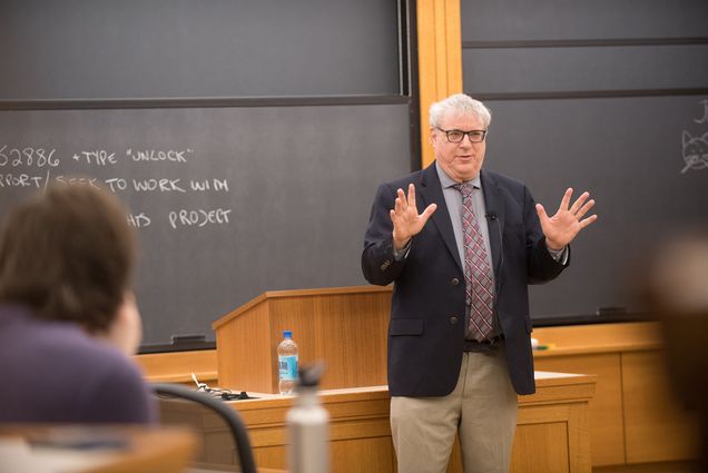 Photo: A picture of a professor teaching at the front of a classroom. His hands are extended as he speaks and there are chalkboards behind him