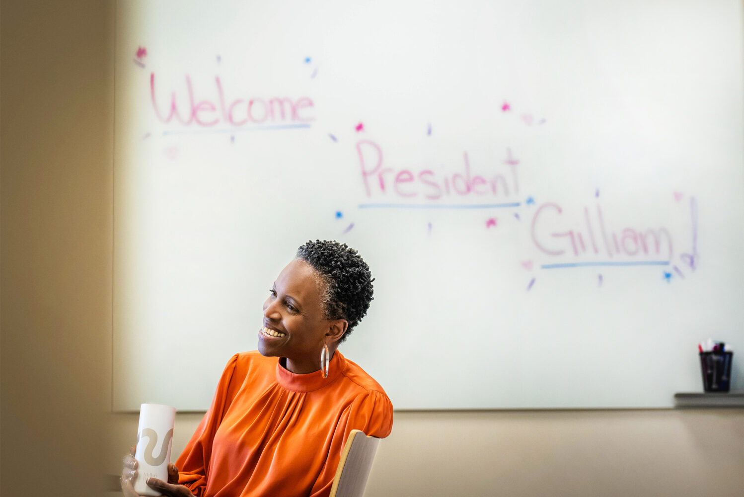 Photo: President Melissa Gilliam sits underneath a whiteboard with a handwritten note reading "Welcome President Gilliam"