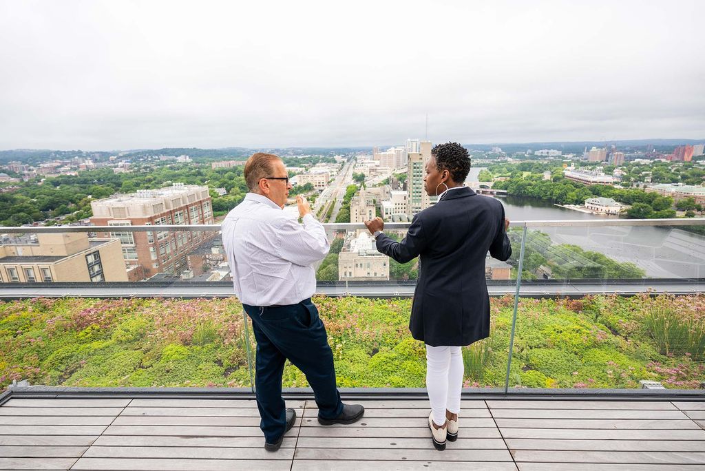 Photo: A middle aged man wearing a collared shirt and a black woman with short hair in a formal outfit look out over a long view of the Commonwealth Ave skyline facing west, away from Boston
