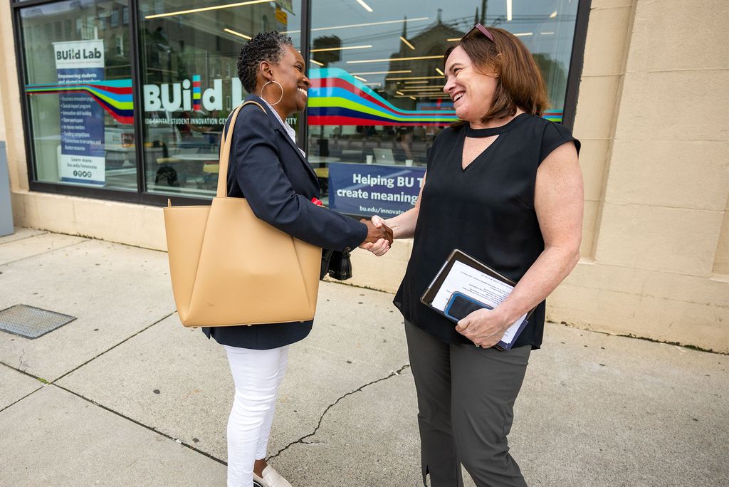 Photo: Two women shake hands, one is a black woman with short hair wearing a colorful tote and the other is a white woman with shoulder length brown hair and glasses on the top of her head