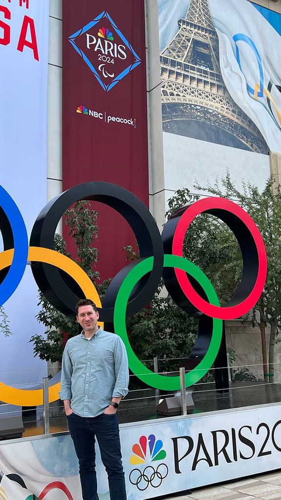 Photo: A man in business casual dress standing in front of the Olympic Rings in Paris for the 2024 Summer Olympic Games