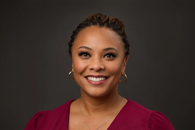 Photo: A black woman with a bright white smile and short dark hair poses for a formal headshot in front of a gray background