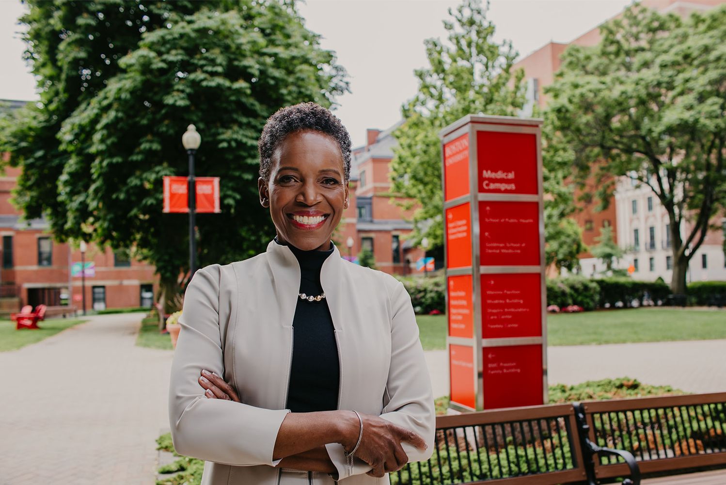 Photo: Boston University's new president Melissa Gilliam, a black woman with short dark hair in a formal outfit stands with arms crossed and a smile in front of a large red Boston University sign, in Boston on a sunny day.