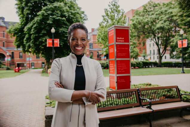 Photo: Boston University's new president Melissa Gilliam, a black woman with short dark hair in a formal outfit stands with arms crossed and a smile in front of a large red Boston University sign, in Boston on a sunny day.