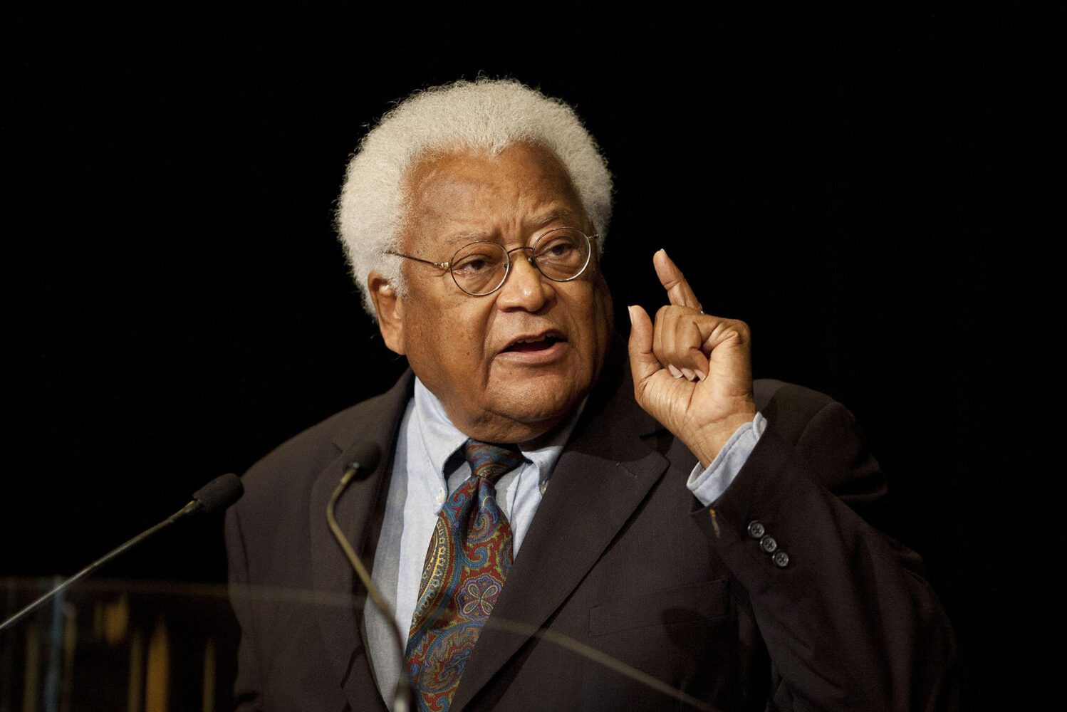 Photo: A picture of James Lawson, an American activist, doing a speech behind a podium while wearing a suit. His hair is white and he is wearing circular glasses
