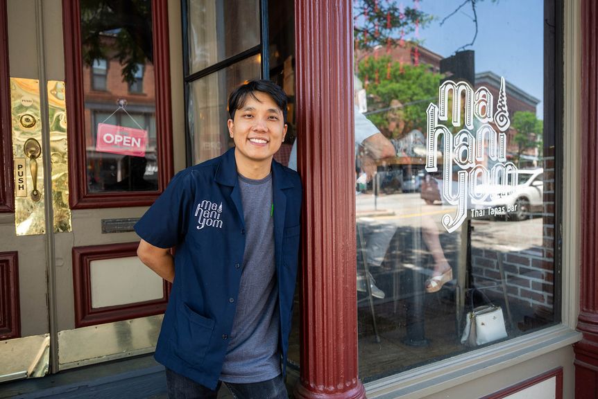 Photo: A young Asian man leans up against his store front. He is wearing a gray shirt and a navy button up.