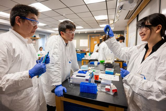 Photo: A picture of students in lab coats, gloves, and protective eyewear gathered around a lab table during an experiment