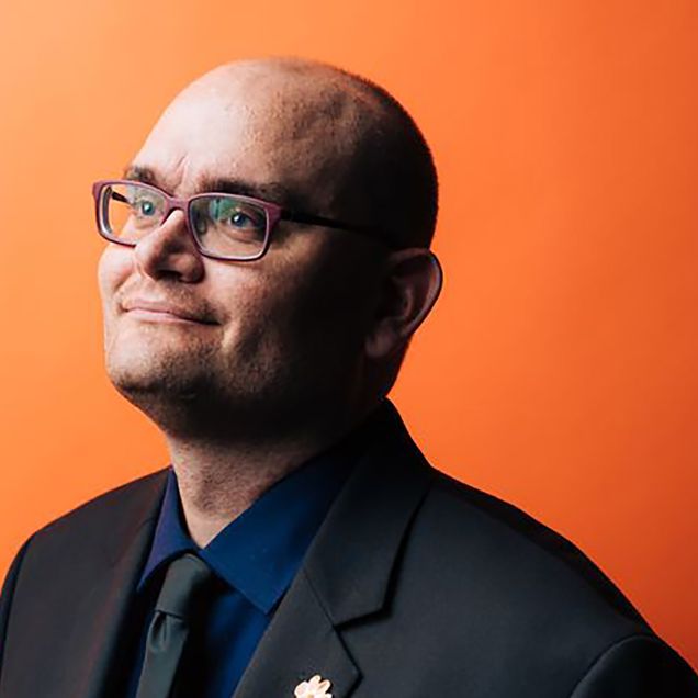 Photo: A bald man looks off to the left with maroon glasses while wearing a dark suit. He stands in front of a orange background.