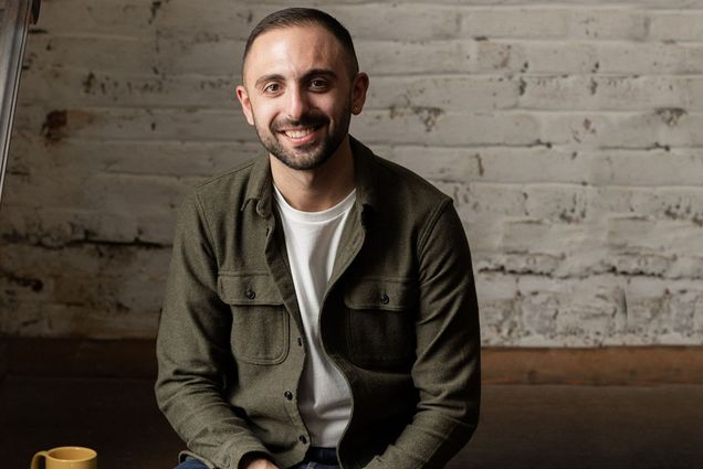 Photo: A picture of a man sitting and smiling ni front of a white brick wall. He is wearing a green jacket, white tshirt, and jeans