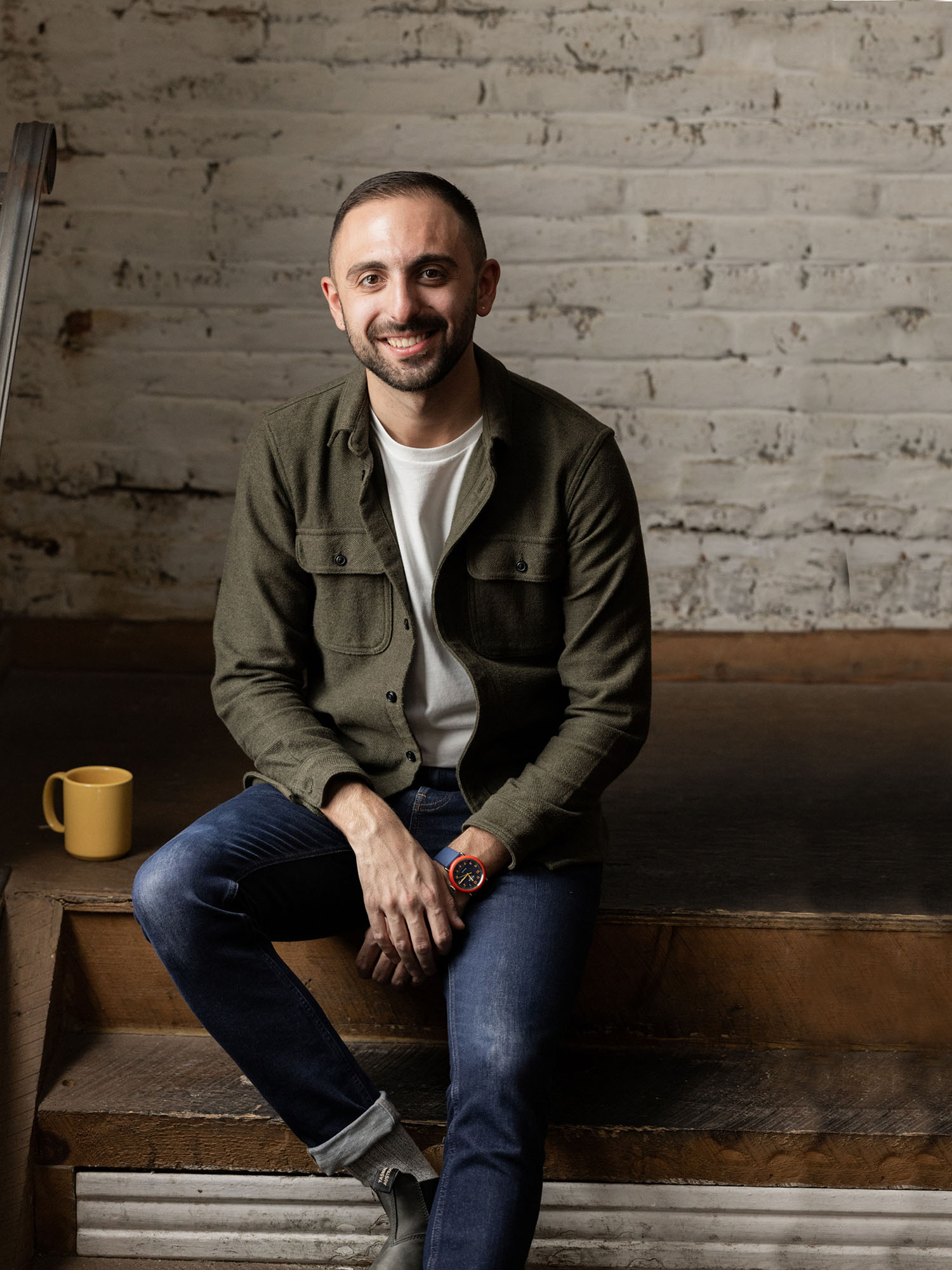 Photo: A picture of a man sitting and smiling ni front of a white brick wall. He is wearing a green jacket, white tshirt, and jeans