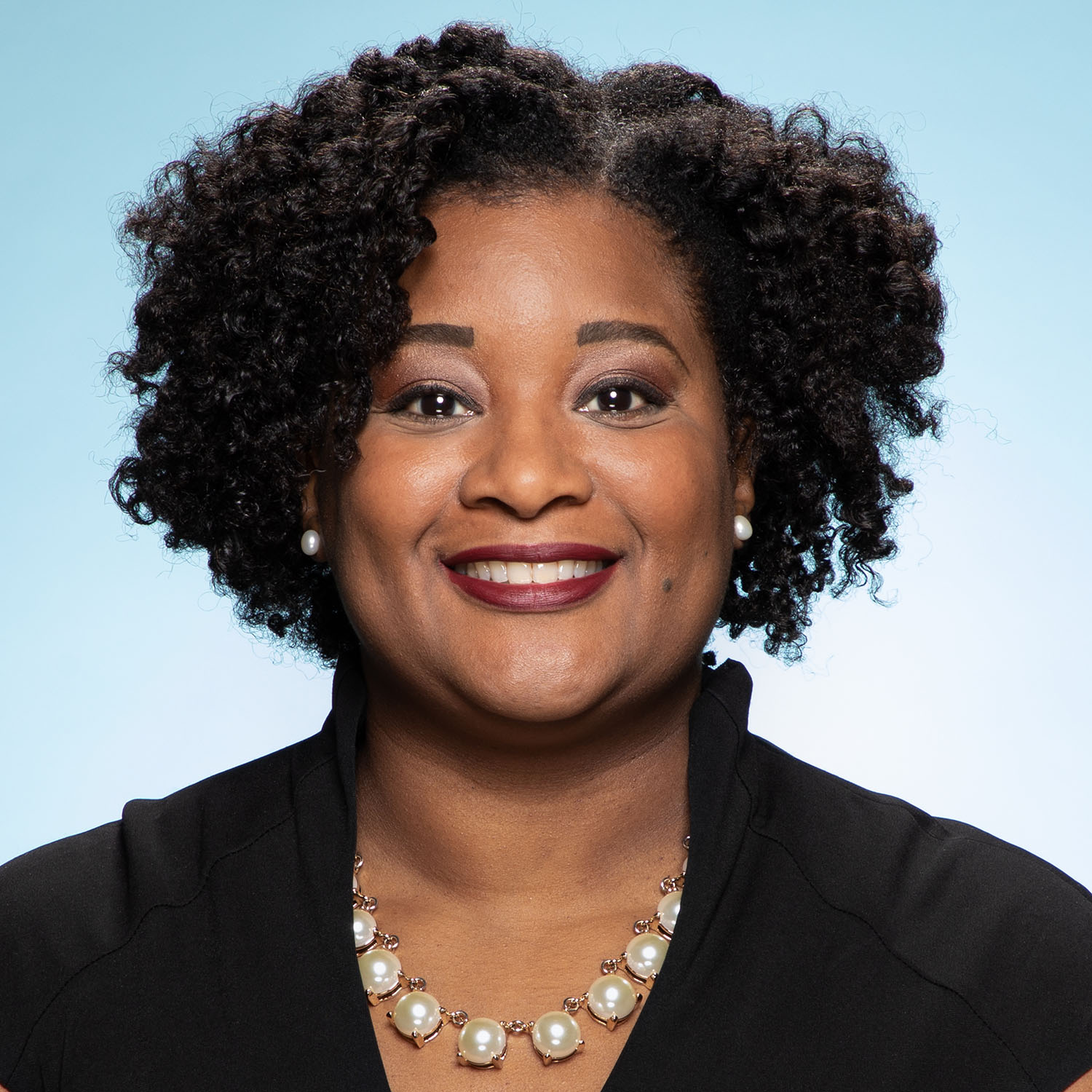 Photo: A headshot of a Black woman wearing a black jacket and pearls. She is smiling in front of a light blue background