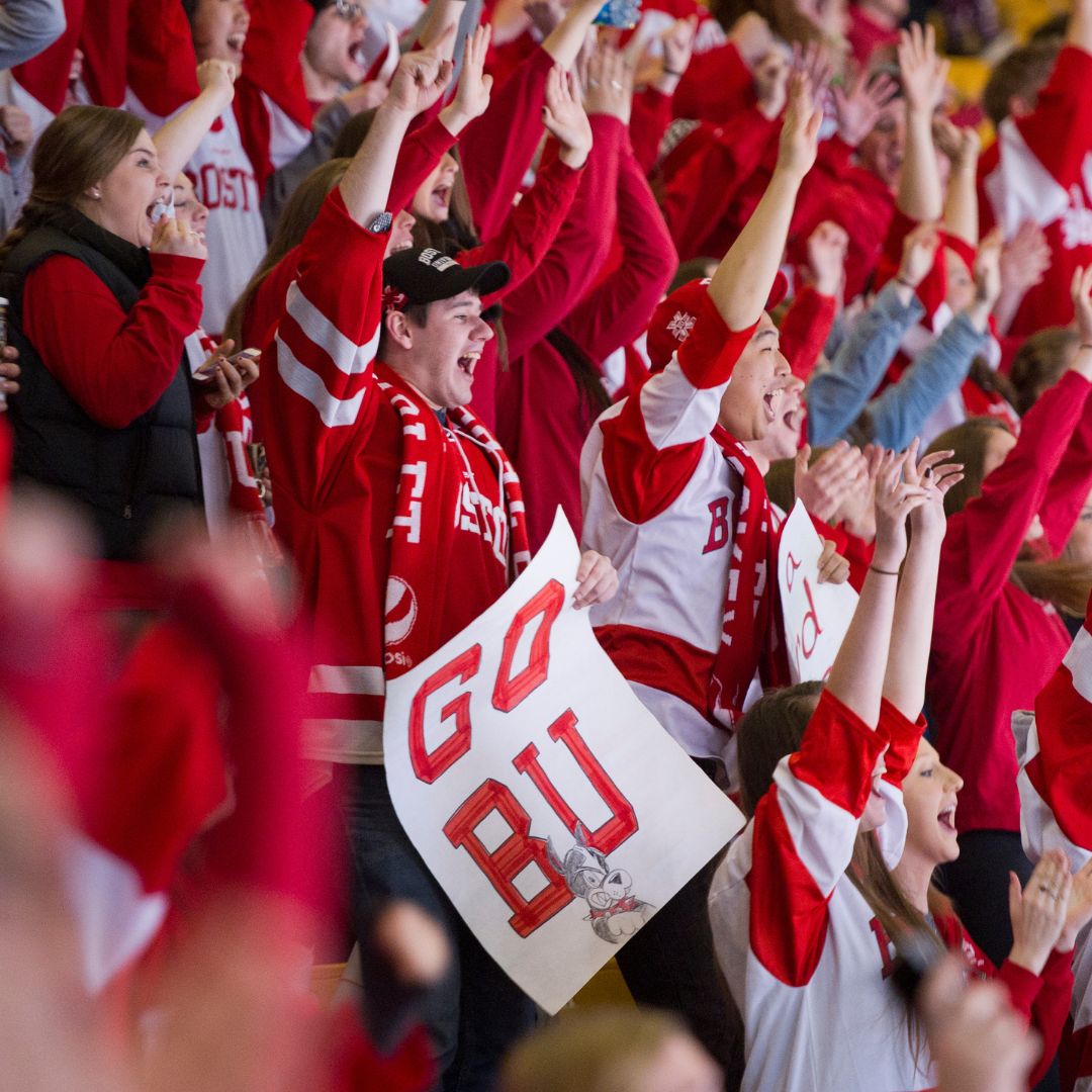 Photo: A group of individuals in a stadium setting, predominantly wearing red attire, with some raising their arms in the air. Faces are obscured by pixelation for privacy. One individual in the foreground holds up a sign that reads ‘GO BU,’ indicating support for Boston University, likely at a sporting event. The atmosphere suggests enthusiasm and team spirit among the attendees.