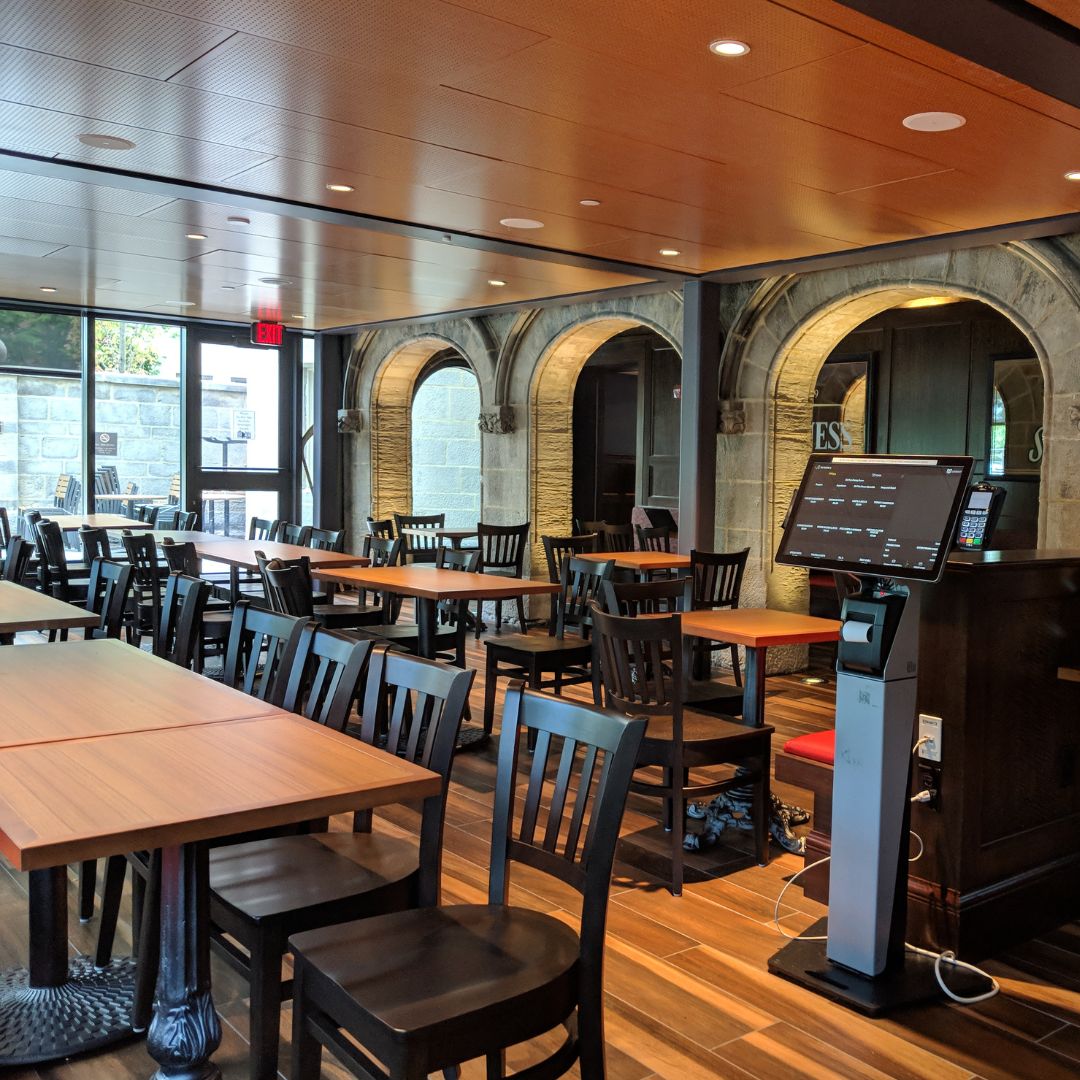 Photo: Interior of Fuller's BU Pub at Boston University with modern and traditional design elements. The space features wooden tables and chairs on a hardwood floor, arched stone doorways on one wall, and large windows offering natural light. A digital ordering kiosk stands in the foreground to the right. The restaurant appears clean and unoccupied, suggesting a photo taken before opening hours or between meal services.