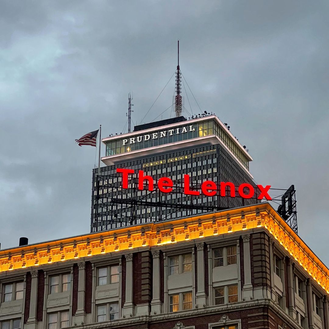 Photo: The image features the top section of two buildings against a cloudy sky at dusk. The foreground shows a building with ornate detailing and warm lighting on its facade, labeled ‘The Lenox.’ Behind it rises a taller building with a flat roof and an array of antennas, prominently displaying the sign ‘PRUDENTIAL’ near its top. An American flag is visible on the right side, mounted on the taller building.