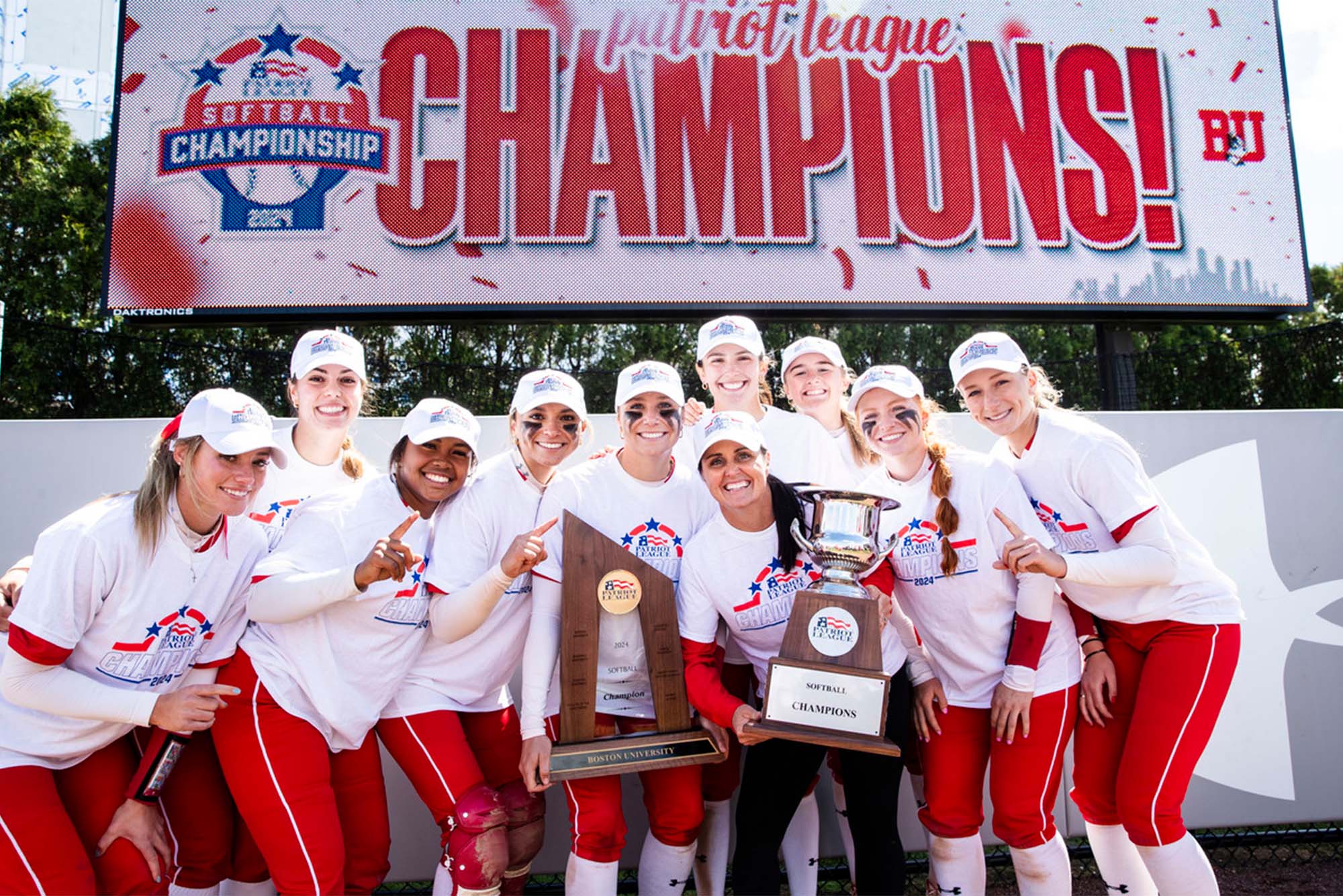 Photo: Boston University's Softball Team holding two large trophies under a banner that reads "Champions"