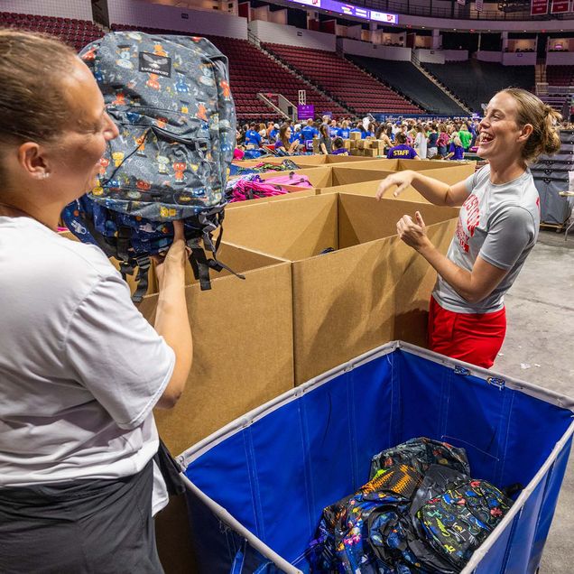 photo: two volunteers standing in a large open room with boxes of backpacks and clothing surrounding them as they sort through