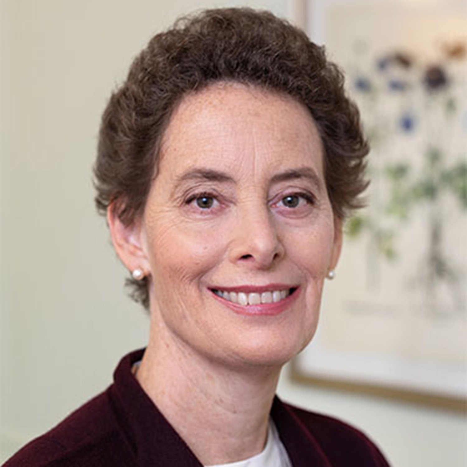 Photo: A picture of a white woman with short, brown hair smiling for a headshot. She is wearing a plum-colored blazer with a white shirt underneath while standing in front of a floral art piece on a white wall.