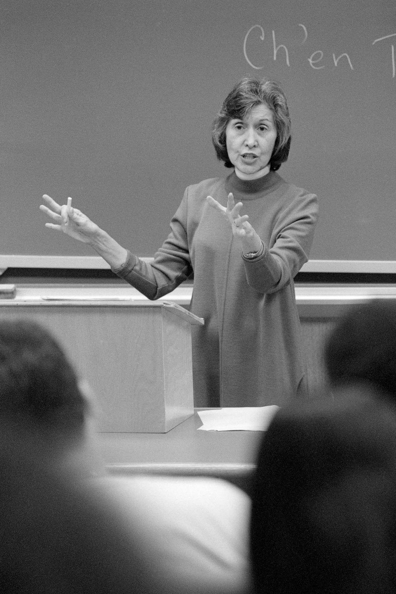 Photo: A black and white photo of a white woman teaching a class. She has short, dark hair and wears a tunic.