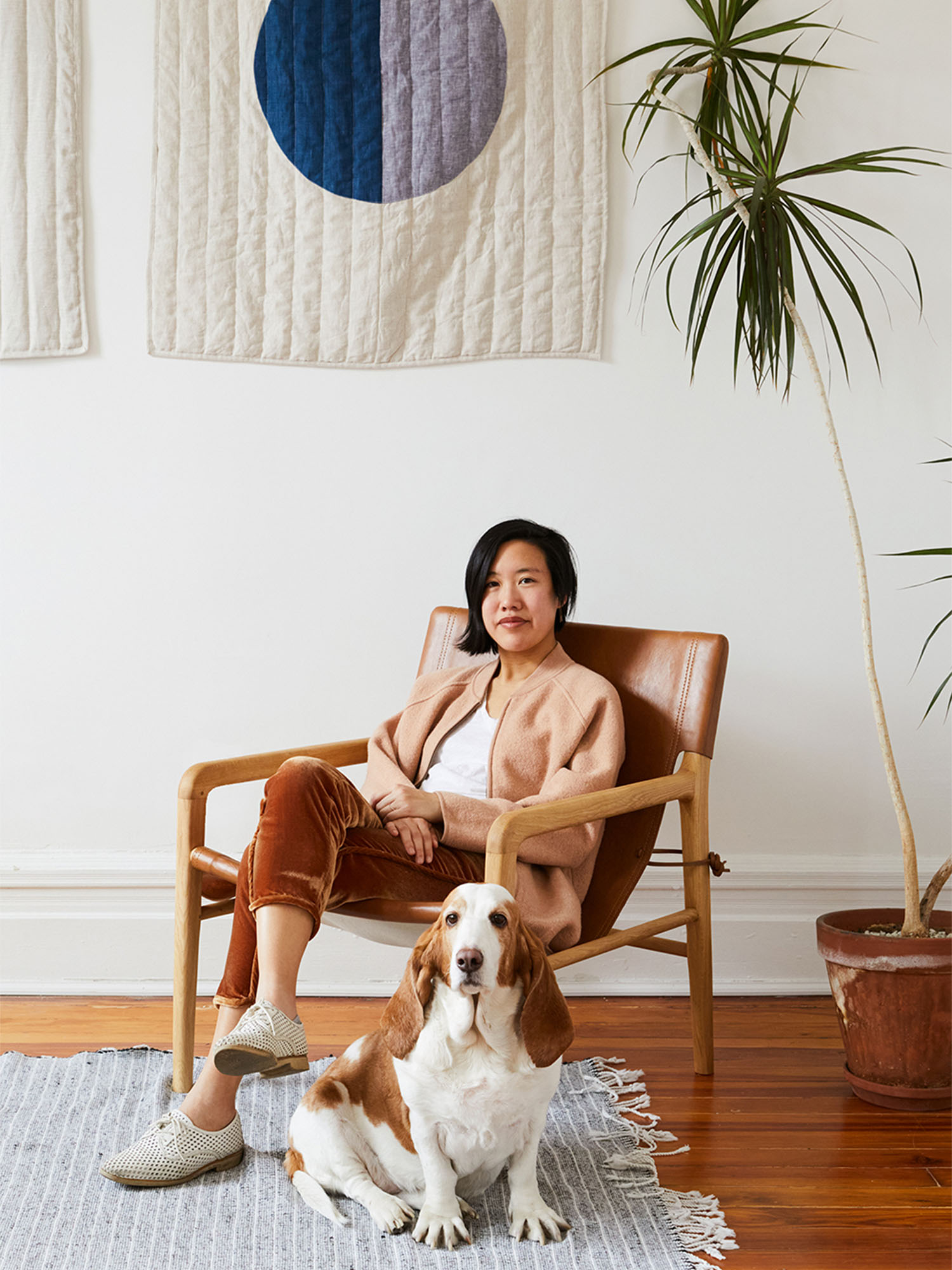 Photo: A woman sits in a mid century modern wooden chair with a basset hound dog in front of the chair. There is a tall plant in the background.