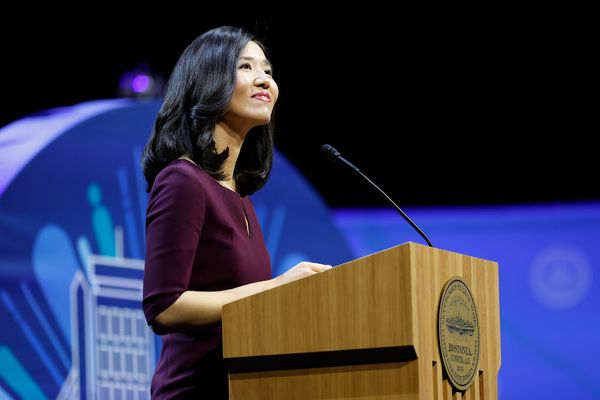 Photo: A picture of Boston Mayor Michelle Wu standing behind a podium. She is wearing a quarter-sleeve burgundy dress and smiling while looking into the crowd