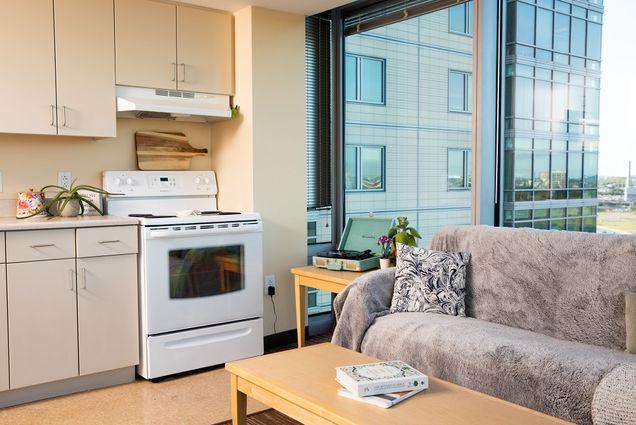 Photo: A picture of a dorm room with a tidy kitchen and living area. The couch has a gray cover over it and there is a seafoam green record player in the corner of the room in front of a large window