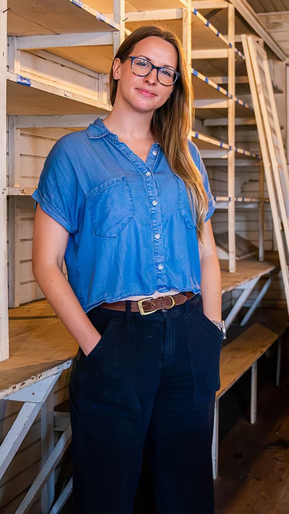 Photo: Mary Snyder, a white woman with shoulder length hair, is a BU PhD student in the American Studies program. She stands in a room with rows of wooden bare shelves.