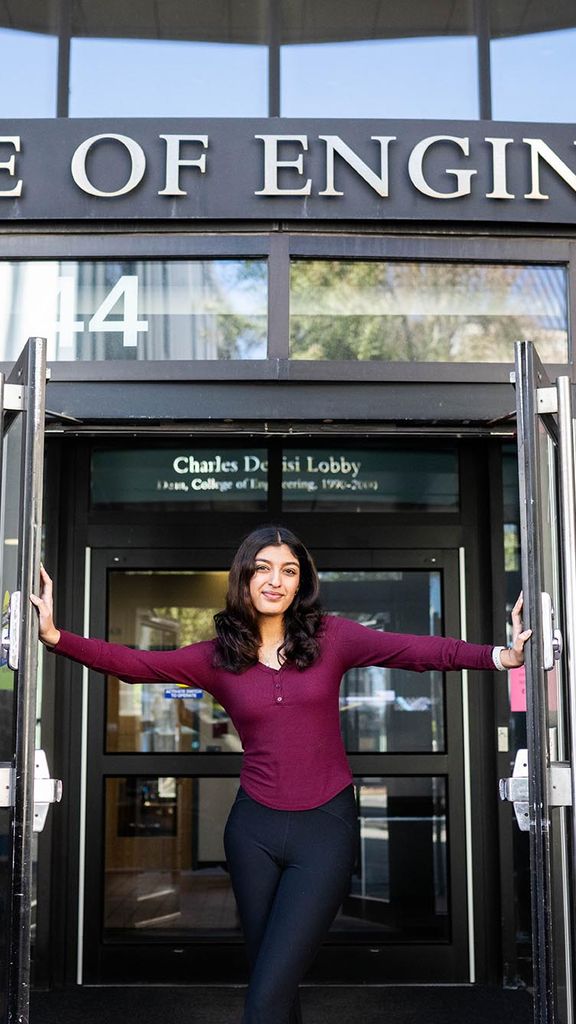 Photo: Aditi Singh, a young woman with black pants and a maroon top, poses in the middle of the doorway to the College of Engineering on BU's campus.