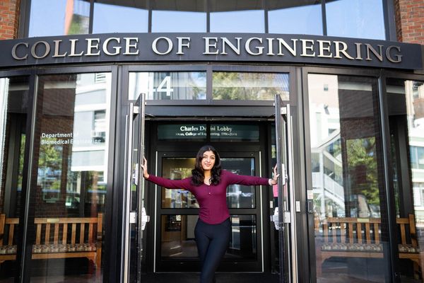 Photo: Aditi Singh, a young woman with black pants and a maroon top, poses in the middle of the doorway to the College of Engineering on BU's campus.