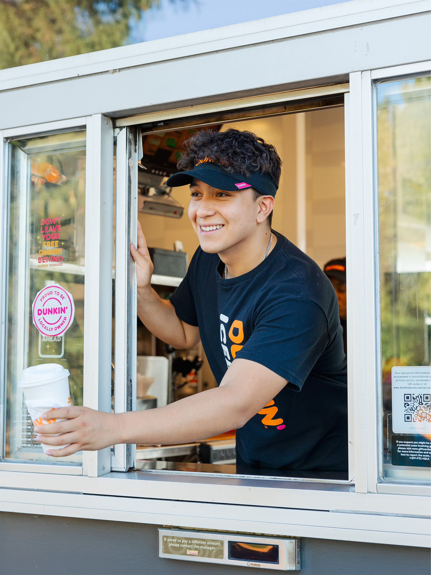 Photo: A young adult male working the drive thru at Dunkin'. He is wearing a black tshirt with the orange Dunkin logo and a visor as he extends a drink from the window