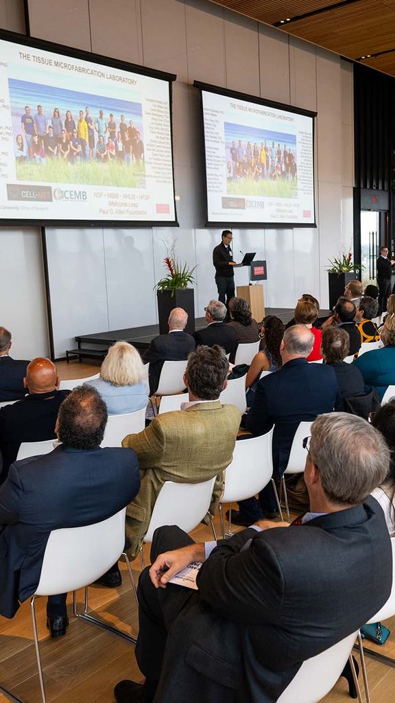 Photo: A large group of people sit watching an individual giving a research presentation in a bright modern space