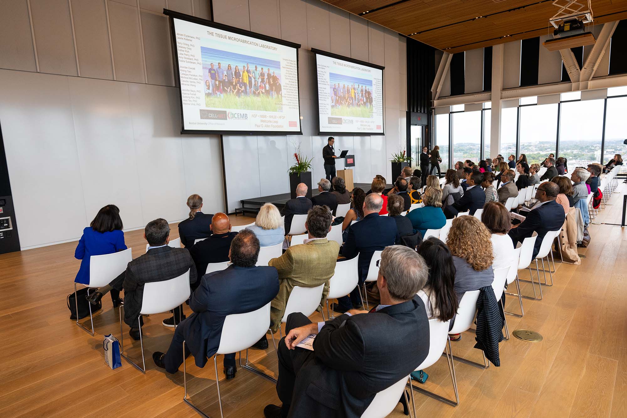 Photo: A large group of people sit watching an individual giving a research presentation in a bright modern space